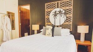 A hotel room with a large bed, two bedside tables with lamps, a wooden headboard with a palm tree design, and a robe rack. - Tamboo Beside the Pointe (Rincon)
