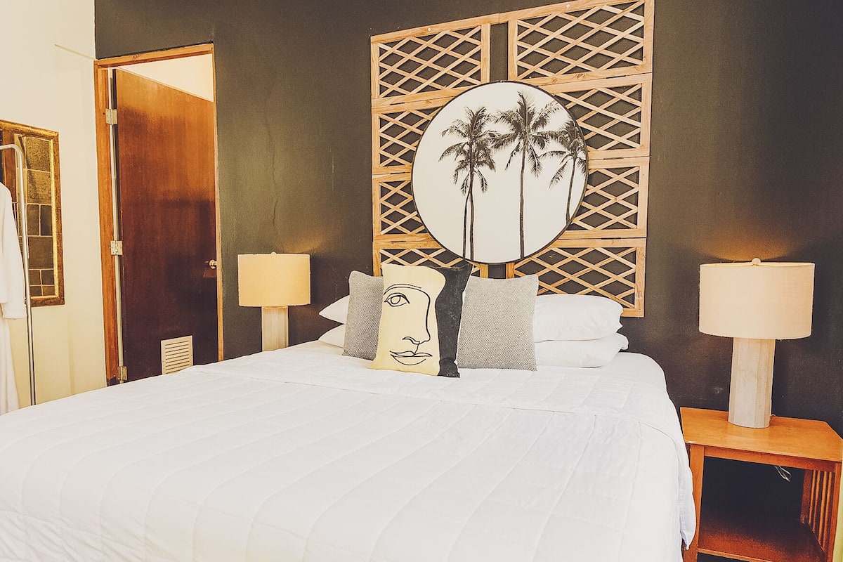 Quarto de hotel com uma cama grande, duas mesas de cabeceira com abajures, uma cabeceira de madeira com design de palmeira e um porta-robes.