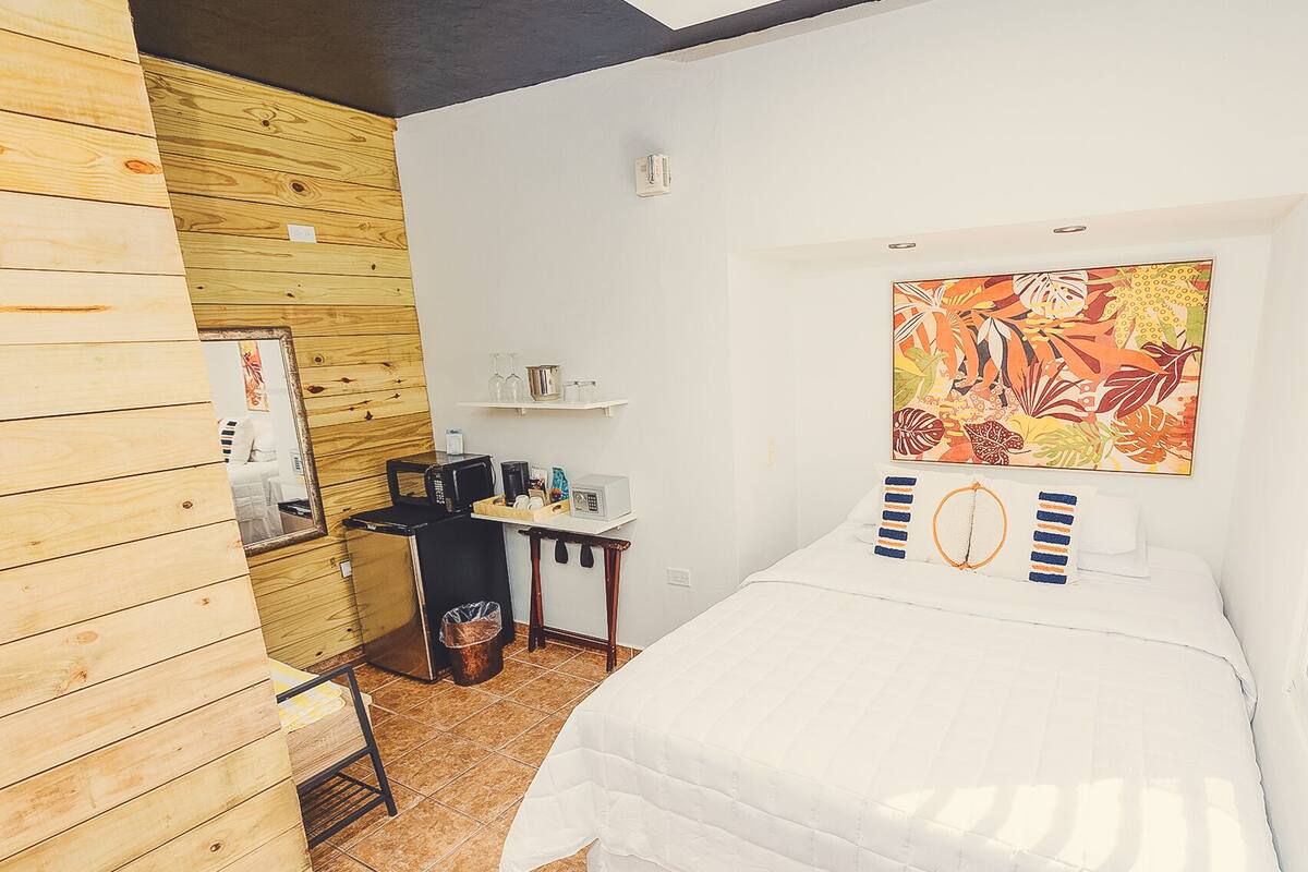 Quarto pequeno e bem organizado, com uma cama, uma escrivaninha com computador e máquina de café, uma estante com copos e um quadro colorido na parede.