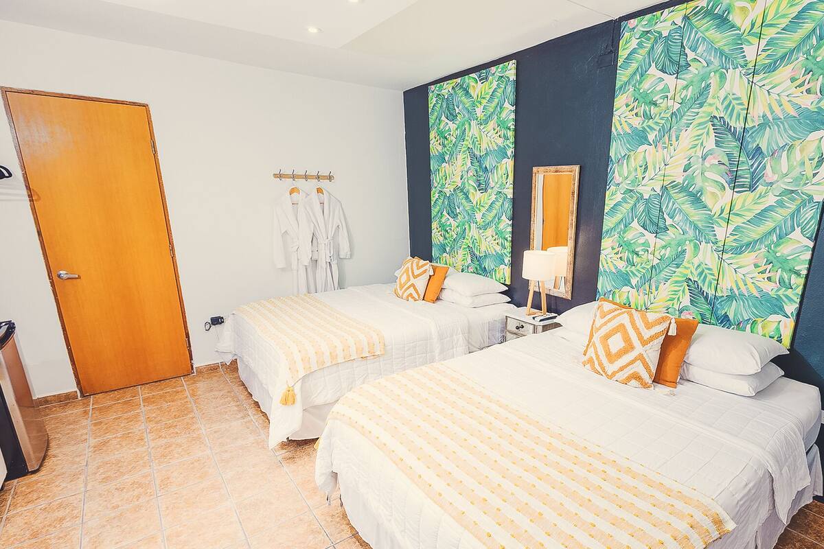 Quarto de hotel com duas camas, porta de madeira, porta-robes e papel de parede com padrão de folhas tropicais.