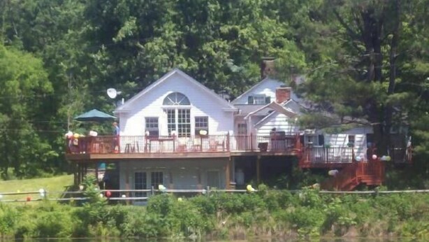A lakeside cabin with a large deck and multiple windows, surrounded by dense forest.