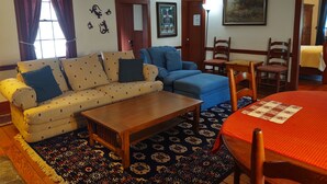 A living room with a sofa, a blue armchair, a wooden coffee table, a patterned rug, and a dining table with a red tablecloth.