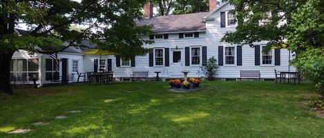 A white house with black shutters, a front porch, and a garden with a birdbath and flowers.