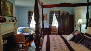 A traditional bedroom with a four-poster bed, a fireplace, a wooden chair, and a desk with a lamp.