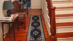 A staircase with wooden steps and a handrail, a wooden bench, a table with magazines, and a rug leading to a room with a bed and a lamp.
