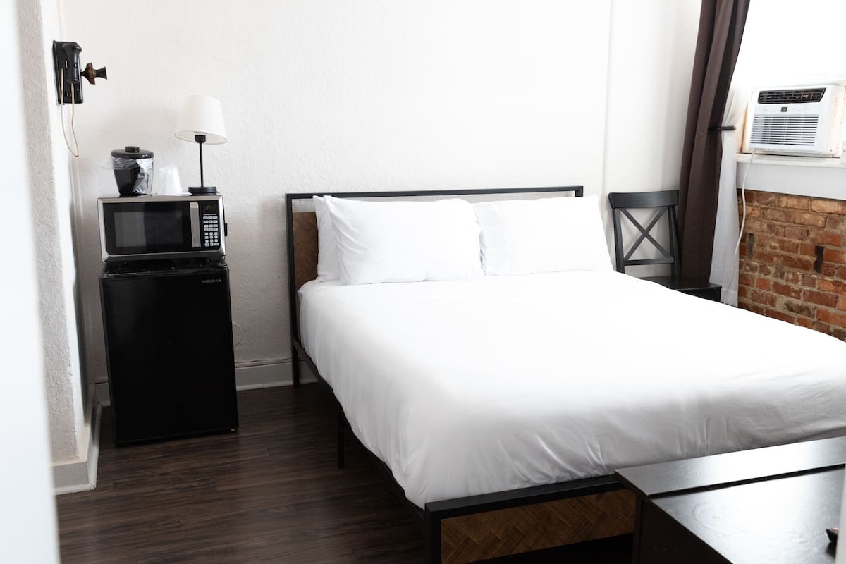 A neatly made bed with white linens, a small black refrigerator, a lamp, and a wall-mounted air conditioner.