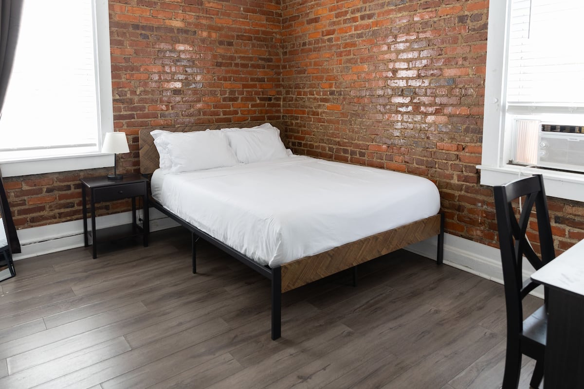 A bedroom with a brick wall, a wooden bed frame, a white mattress, a nightstand, a chair, and a window with blinds.