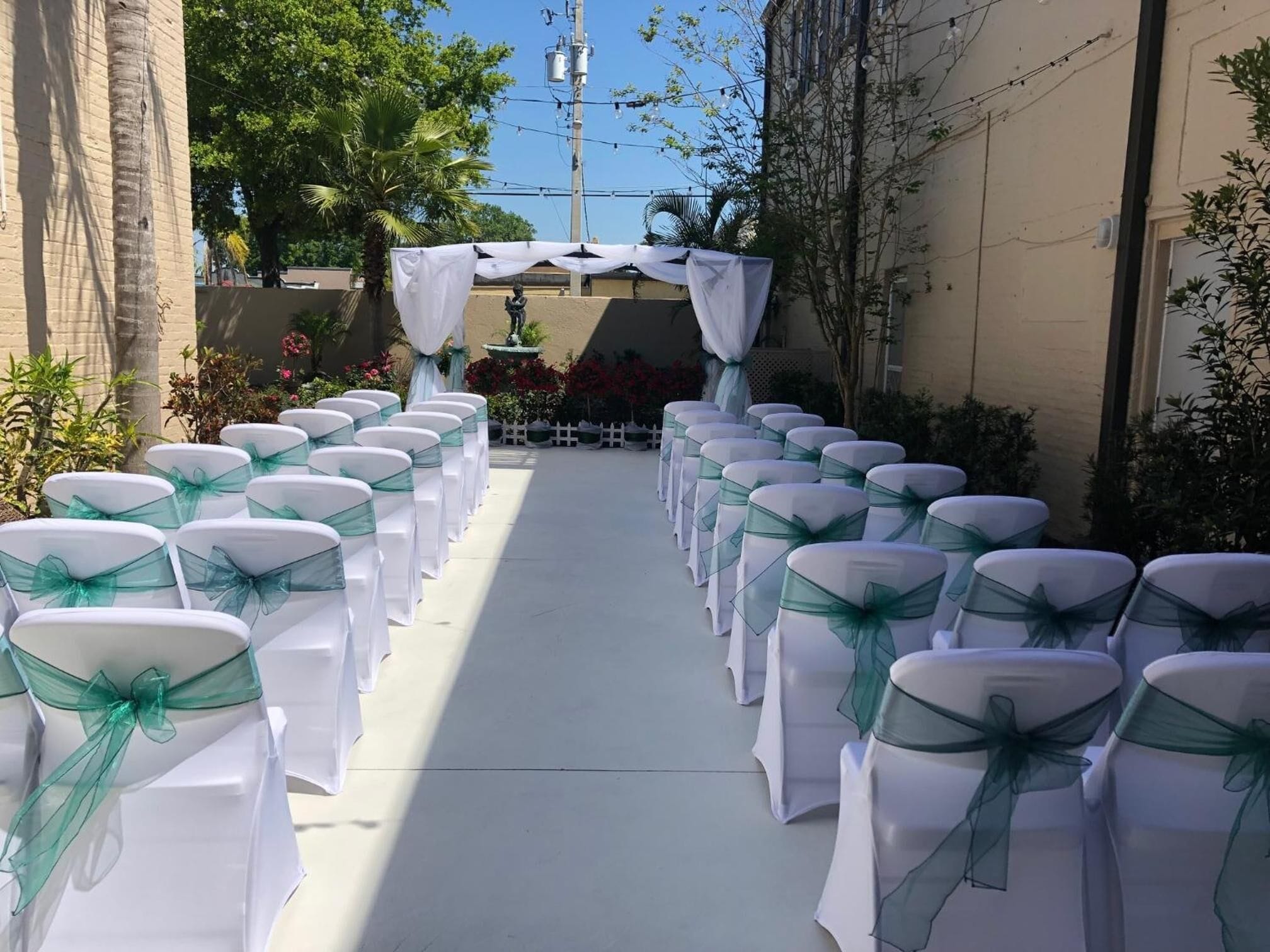A pathway lined with white chairs with green sashes, leading to a white archway with a fountain.