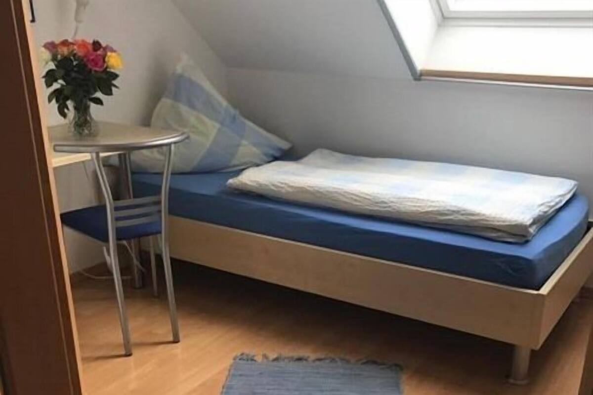 Een kleine kamer met een enkel bed, een schuine wand, een tafeltje met een stoel en een raam voor natuurlijk licht.