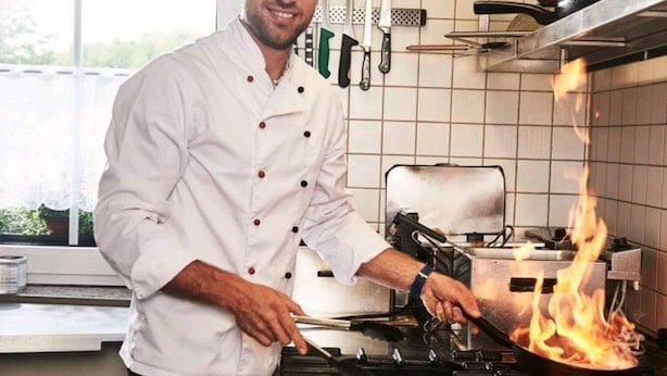 Un chef, vêtu d’un uniforme blanc, cuisine à l’aide d’une poêle sur une cuisinière dans une cuisine aux murs carrelés, où divers ustensiles de cuisine sont suspendus au-dessus.