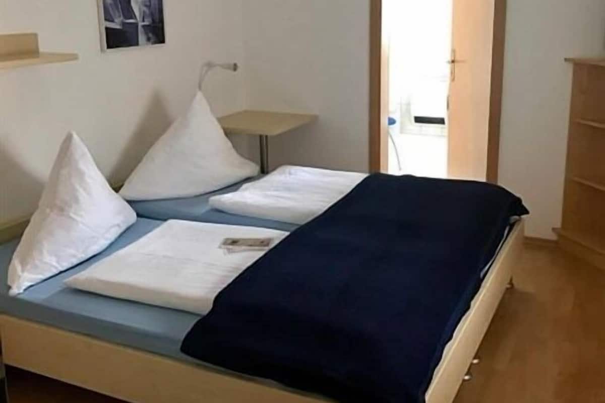 Een bed met witte kussens en een donkerblauwe deken, een klein nachtkastje en een wandlamp.