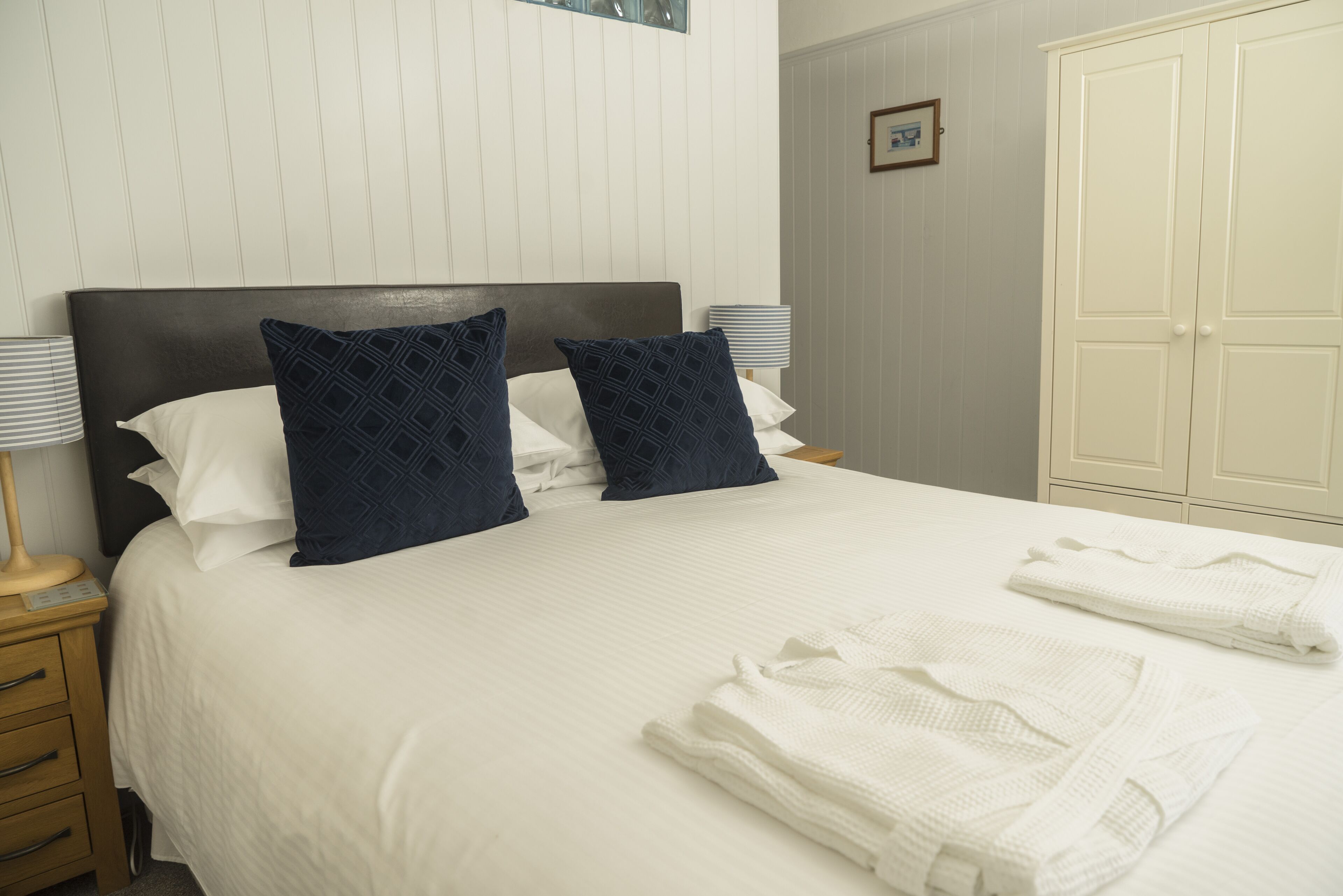Una cama bien tendida con ropa blanca y dos almohadas oscuras de estampado azul, una lámpara de noche y batas blancas dobladas sobre la cama.