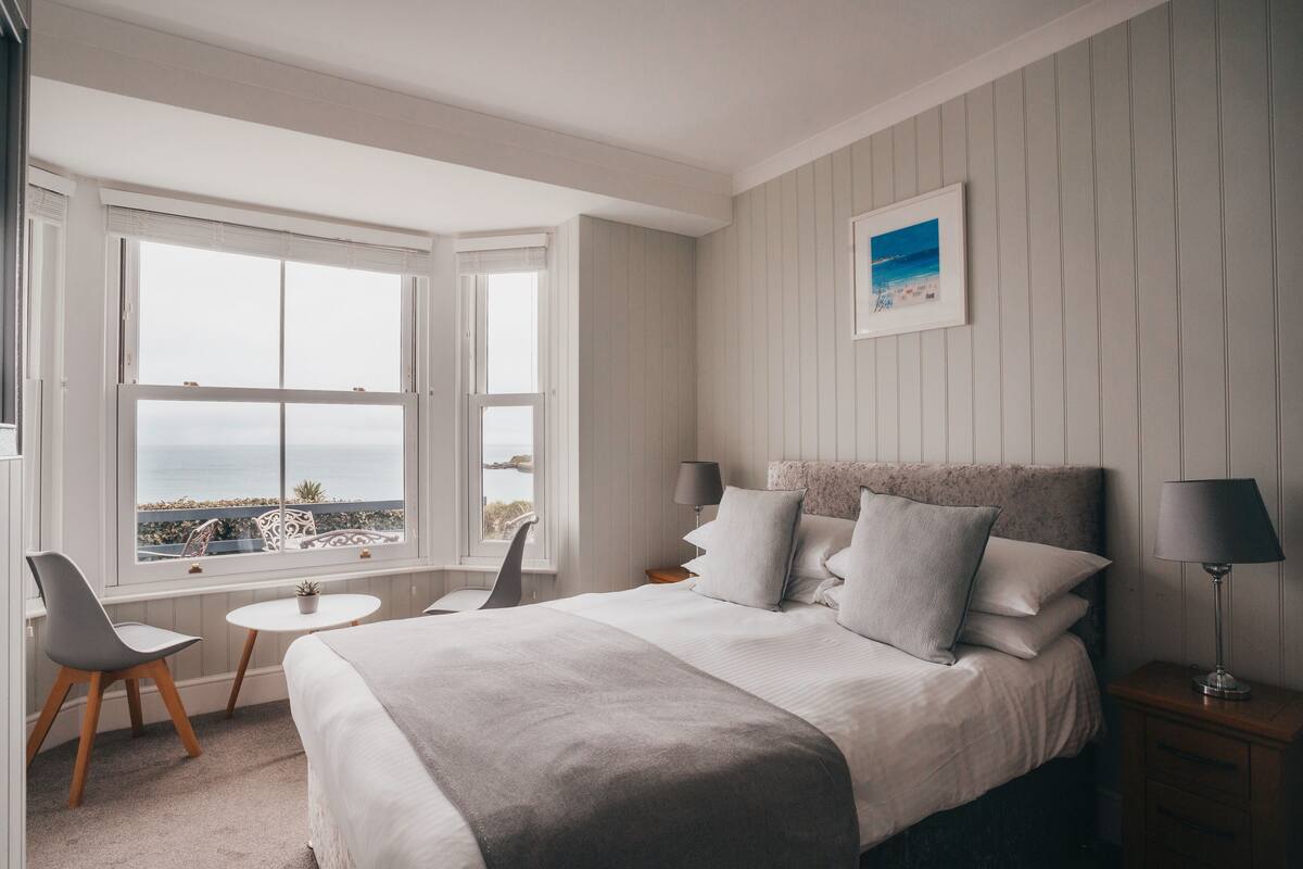 A bedroom with a large bed, a desk, and a chair. There is a window with a view of the ocean.