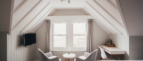 Een slaapkamer met een schuine plafond, twee witte stoelen, een klein rond tafeltje, een bed, een bureau met stoel en een aan de muur bevestigde televisie.