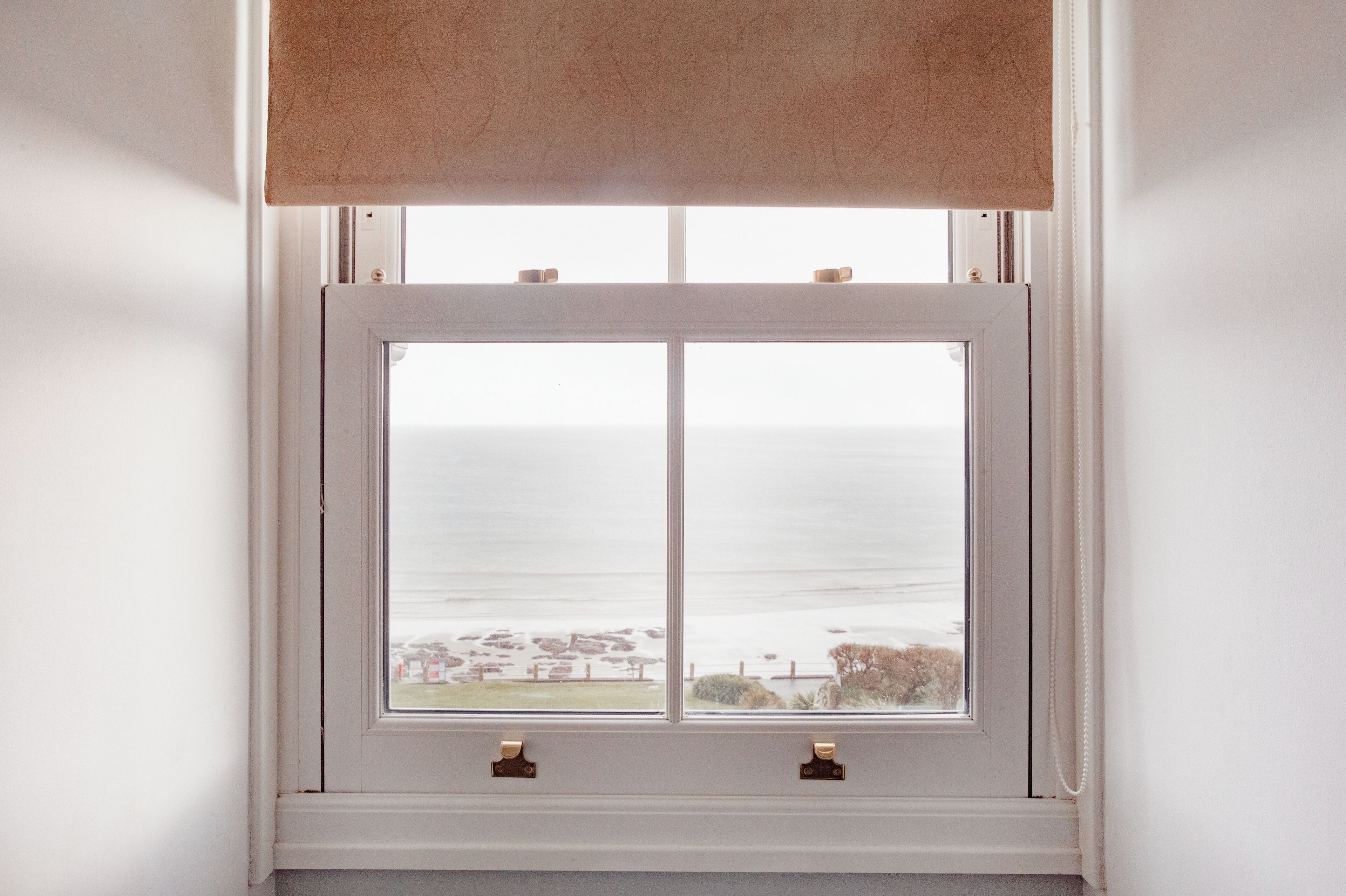 A window with a white frame and two handles, overlooking a coastal view with a beach and ocean.