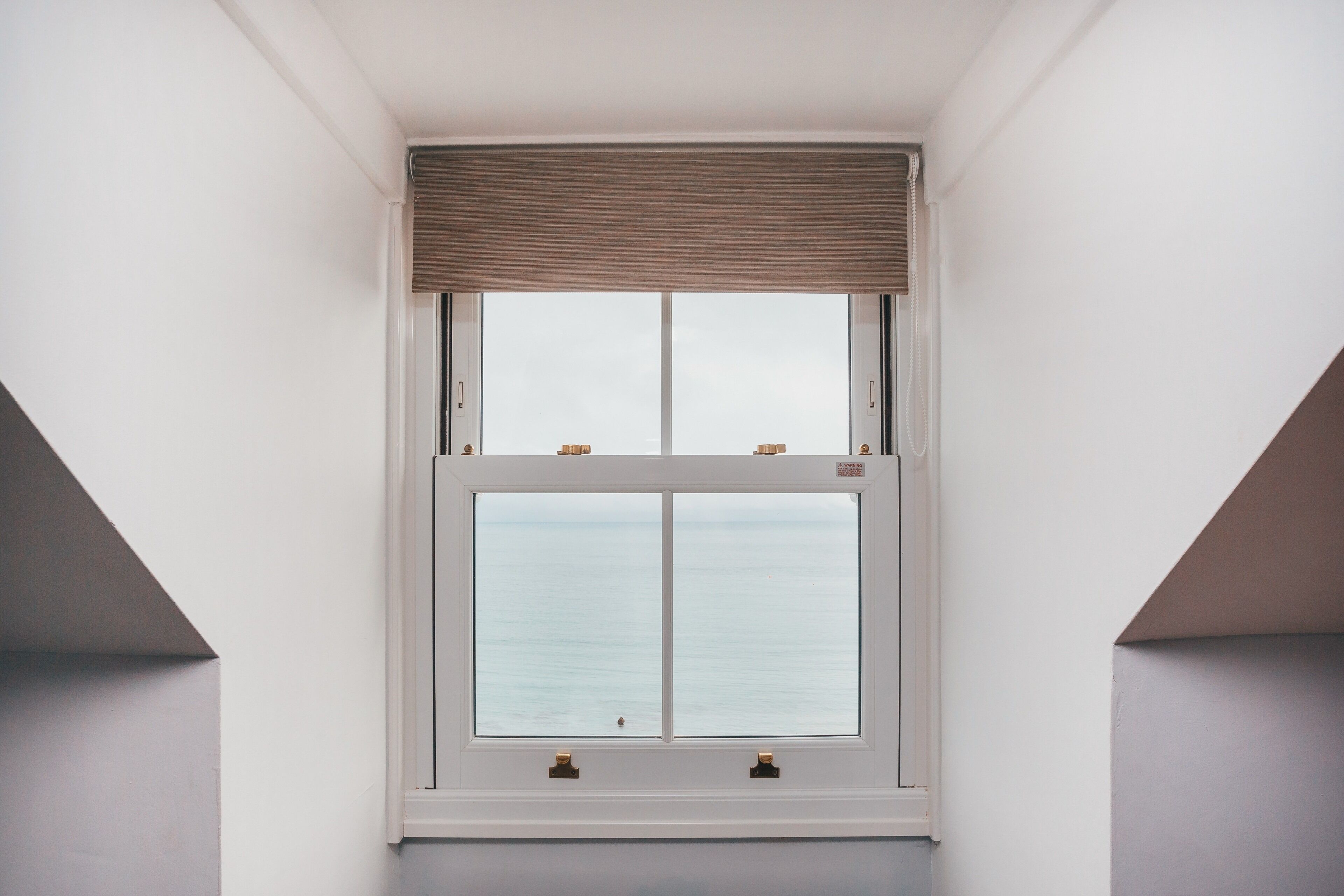 A window with a view of the sea, white walls, and a brown window blind.