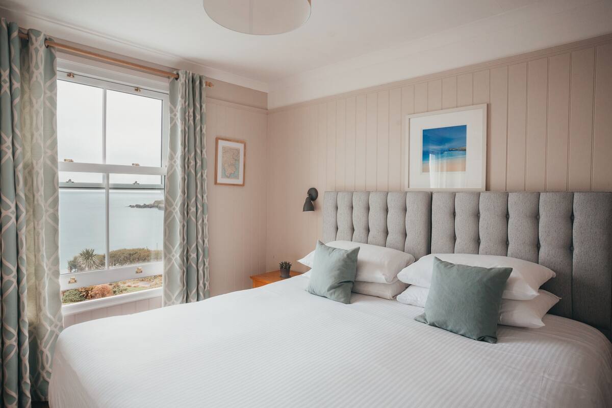 A neatly made bed with pillows, a bedside table with a lamp, a framed picture on the wall, and a window with curtains.