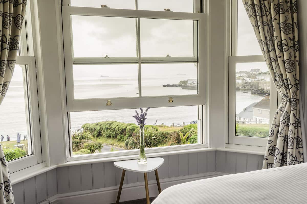 A room with a large window overlooking a coastal view, a small white table with a vase, and a bed with white bedding.