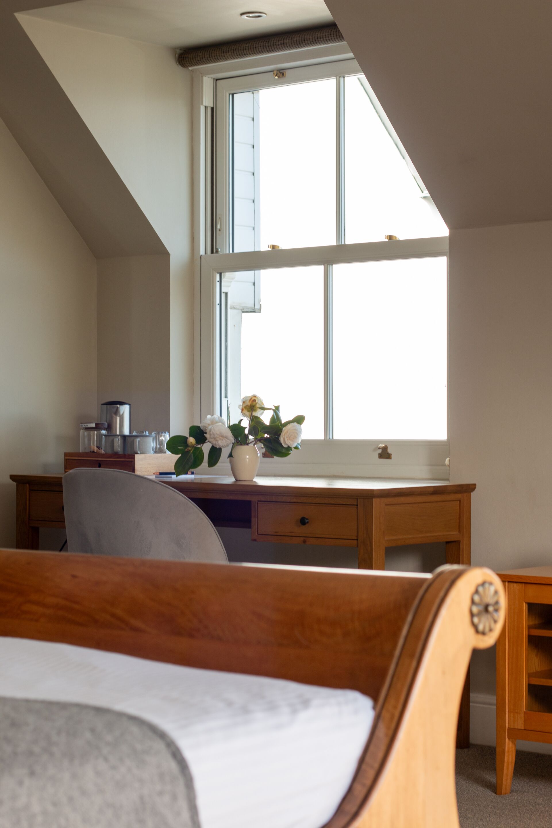 A bedroom with a sloped ceiling, a wooden desk, a chair, a bedside table, and a window with a view.