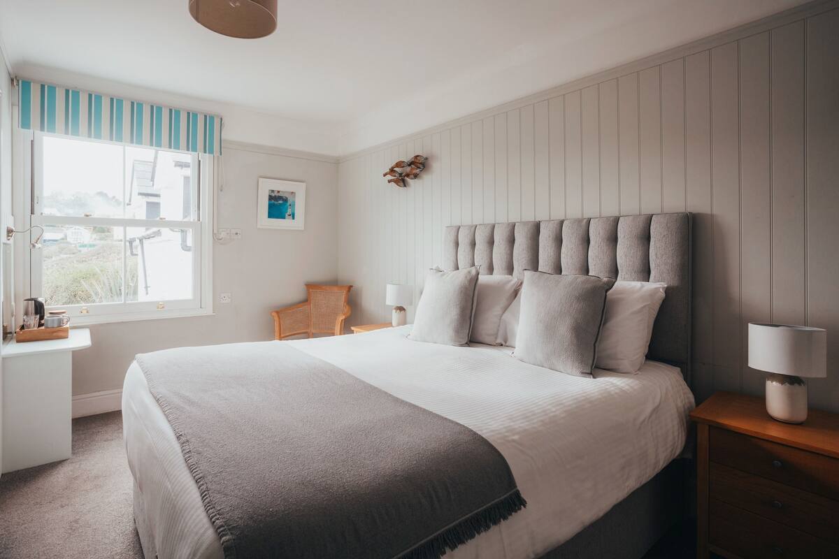 A bedroom with a large bed, a bedside table with a lamp, a window with a view, and a small desk with a chair.