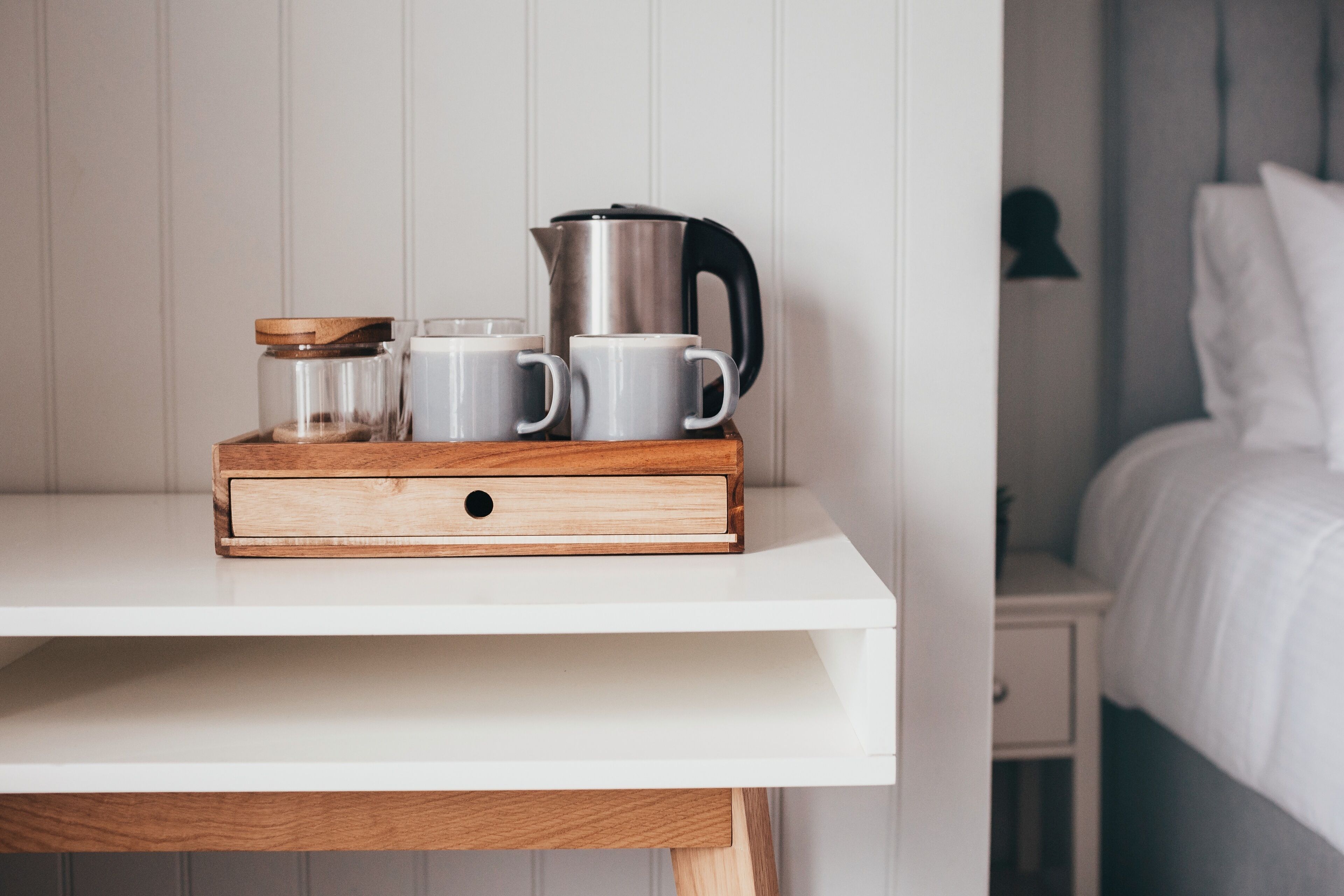 A white bedside table with a wooden tray holding a kettle, two white mugs, and a glass jar.
