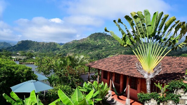 A red-roofed building with a hammock and a large banana plant in the foreground, surrounded by lush greenery and hills in the background. - La Casa de Cafe (Copan Ruinas)