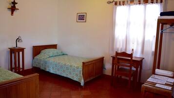 Un dormitorio con cama de madera, una silla de madera, una mesita con papeles, una lámpara de pared y una ventana con cortinas.