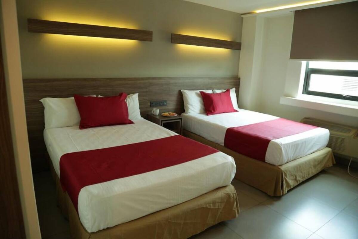 A hotel room with two beds, each with a red and white bedspread, wooden headboards, and a nightstand with a lamp.