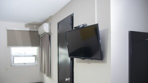 Una habitación con televisor de pantalla plana en la pared, una ventana con cortinas, una puerta y un cubo de basura pequeño.