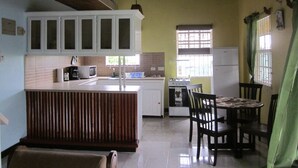 A compact kitchen with white cabinets, a central island, a dining table with chairs, a stove, and a refrigerator. - Buttercup Cottage Apartments (Arnos Vale)