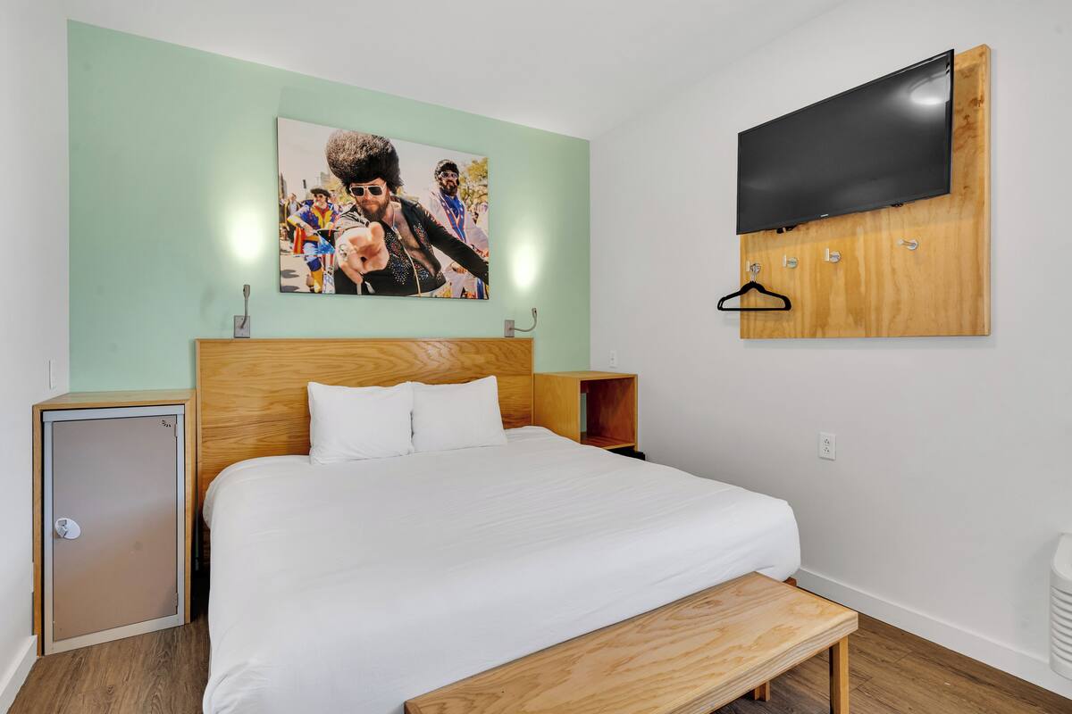 Un dormitorio con cama, cabecera de madera, televisor montado en la pared, una pequeña alacena y un banco.