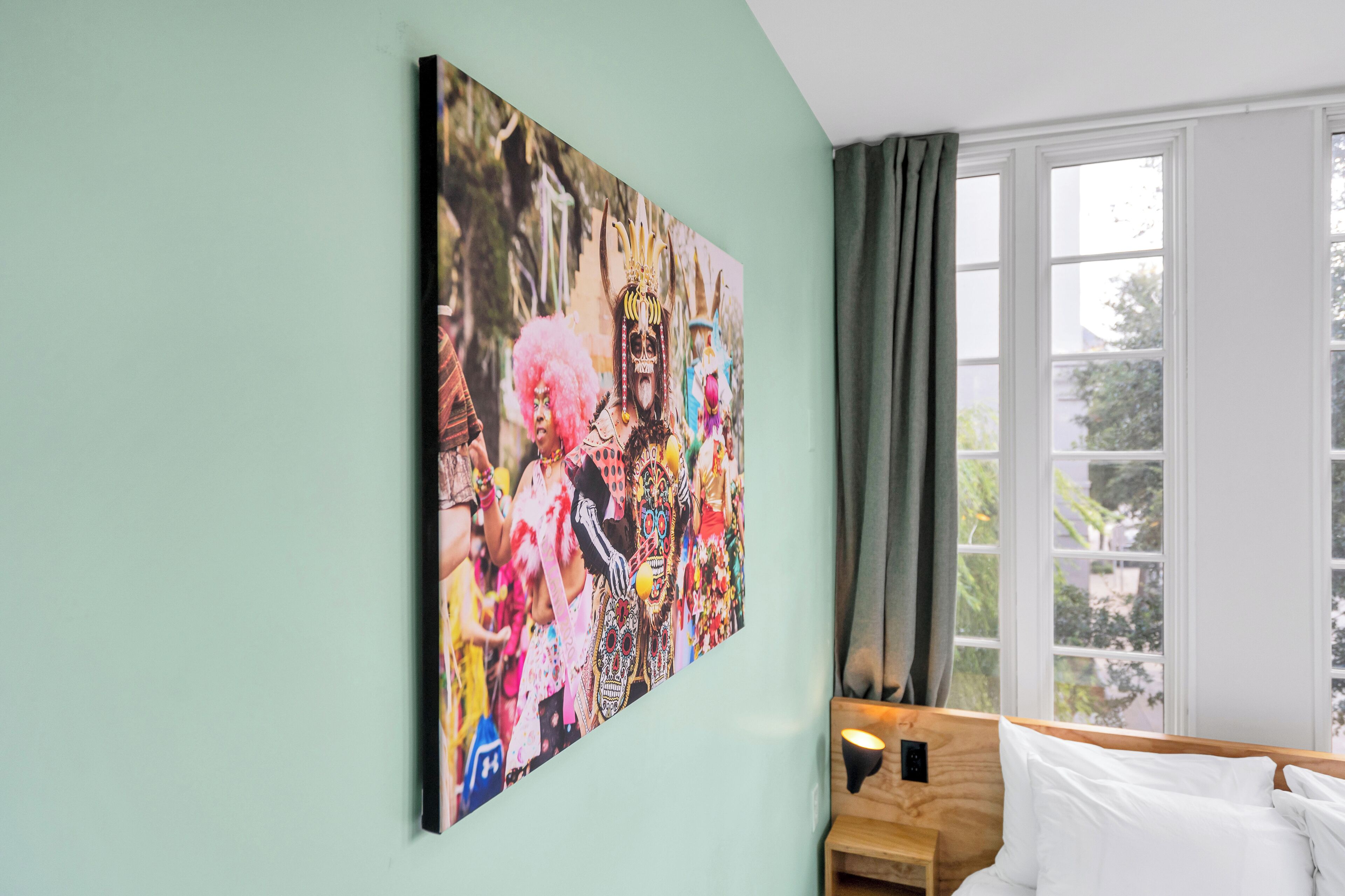 Une chambre avec un grand tableau coloré au mur, un lit avec du linge de maison blanc, une table de chevet en bois et une fenêtre avec des rideaux.