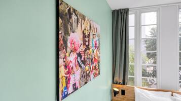 Une chambre avec un grand tableau coloré au mur, un lit avec du linge de maison blanc, une table de chevet en bois et une fenêtre avec des rideaux.