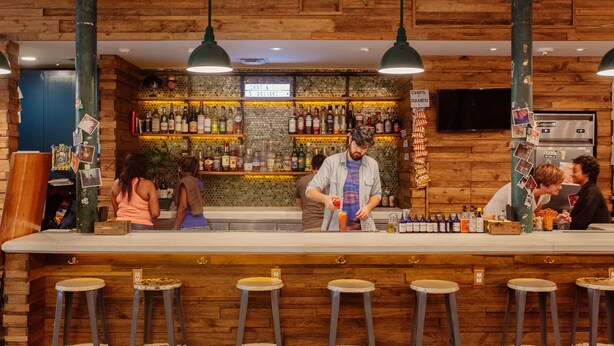 A bar with wooden walls, a counter, and stools. There are bottles and a sign indicating prices. - The Quisby Hotel (New Orleans)