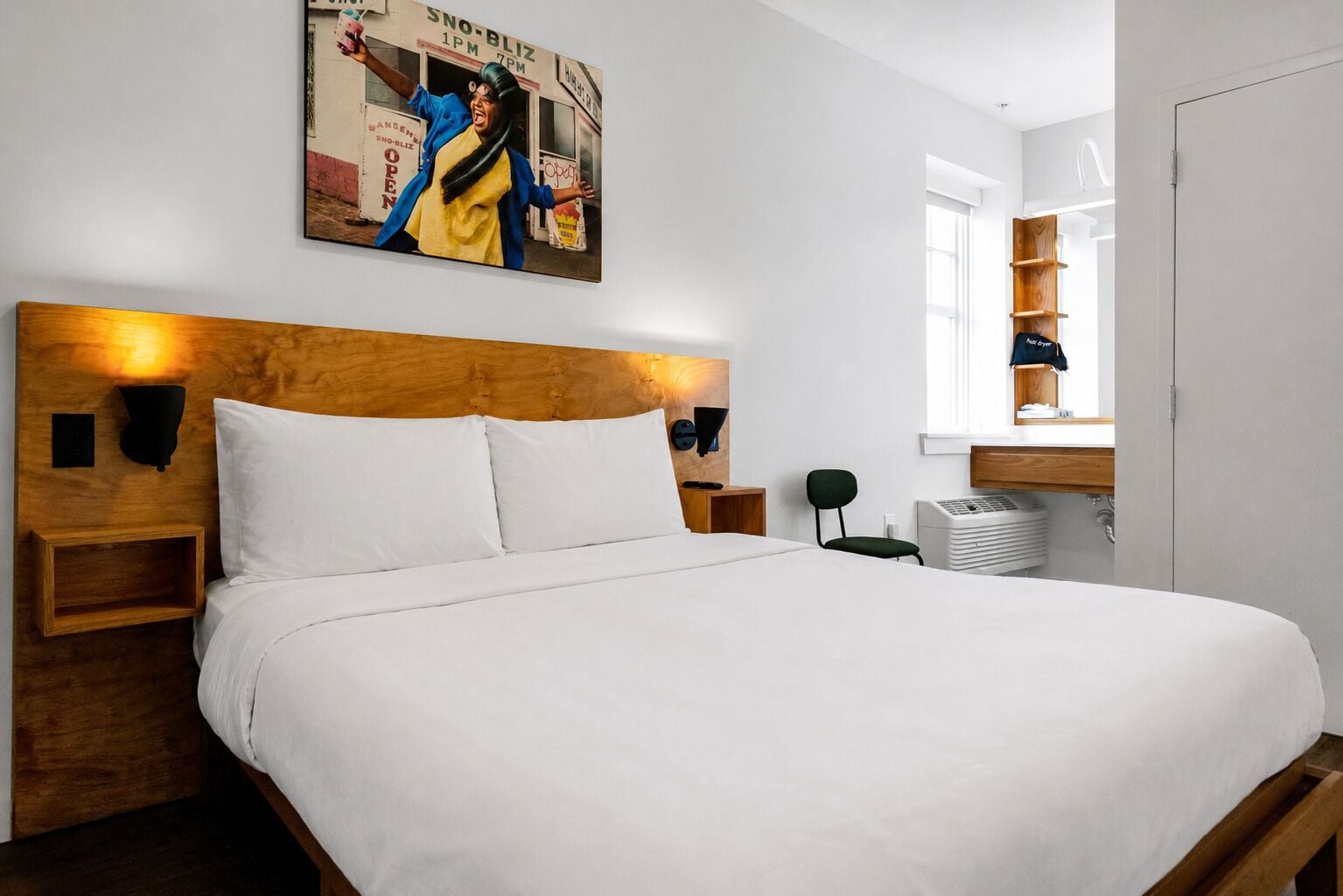 Une chambre d’hôtel avec un grand lit, une tête de lit en bois, deux tables de chevet, une petite table, une lampe fixée au mur, une fenêtre donnant sur un immeuble et une étagère avec une serviette et une chemise.