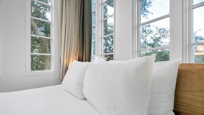 A neatly made bed with white linens and pillows, large windows with curtains, and a view of trees outside. - The Quisby Hotel (New Orleans)