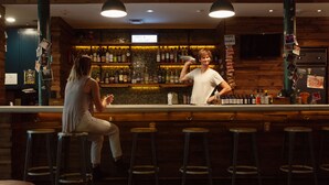 A bar with stools, a bartender, and a shelf with bottles. - The Quisby Hotel (New Orleans)