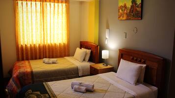 Quarto de hotel com duas camas, um quadro na parede, uma mesa de cabeceira com abajur e uma janela com cortinas.