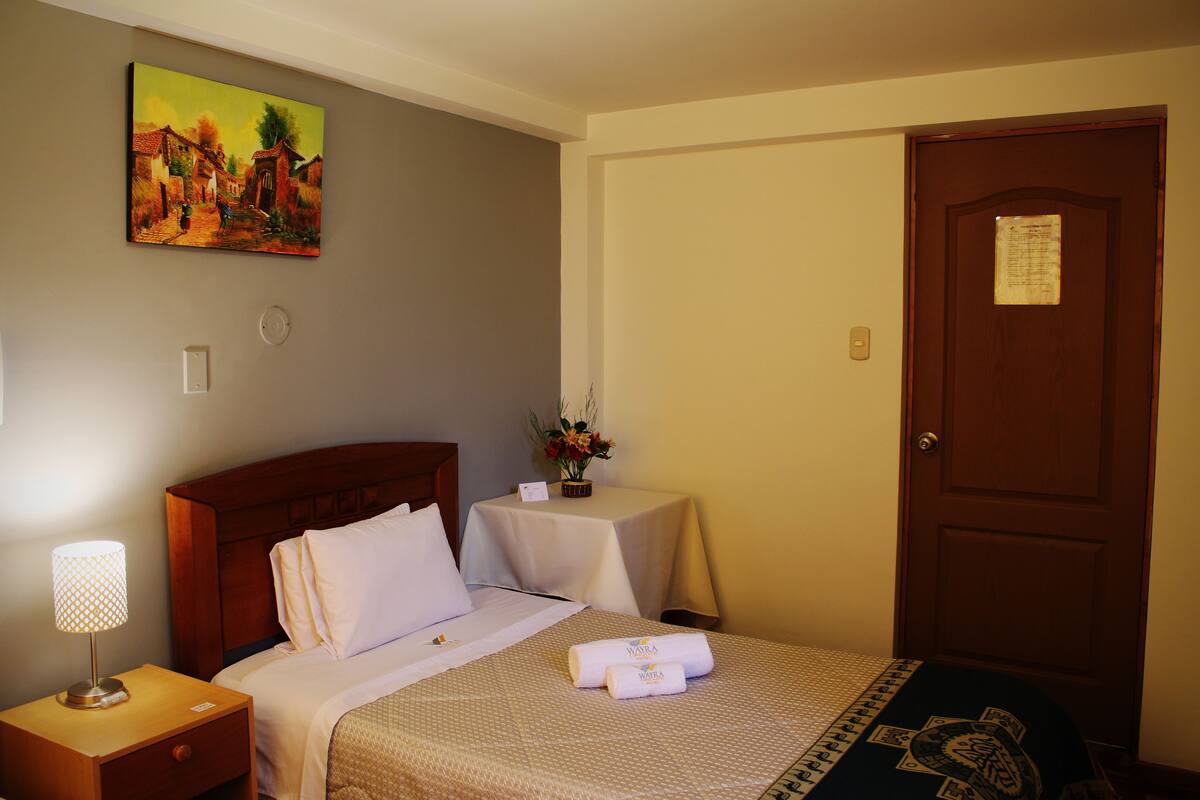 Une chambre d’hôtel avec un lit, une table de chevet avec une lampe, une petite table avec un vase de fleurs et une porte avec une pancarte.