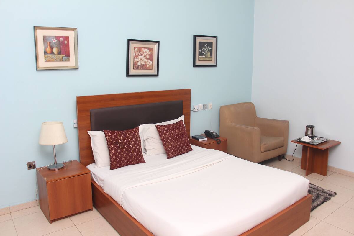 A neatly arranged bedroom with a bed, a nightstand, a chair, and a small table with a phone and a tray.