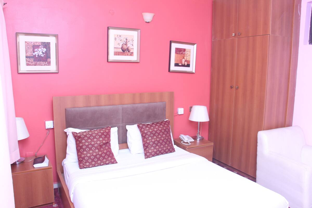 A hotel room with a pink wall, a wooden wardrobe, a bed with white bedding and patterned pillows, a bedside table with a lamp, and a phone.
