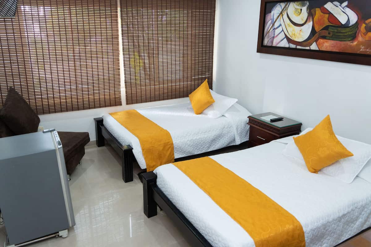 Ein Hotelzimmer mit zwei Betten, beide mit weißer Bettwäsche und gelben Akzenten. An der Wand hängt ein Bild, ein Fenster mit Jalousien und ein kleiner Tisch mit einer Fernbedienung.