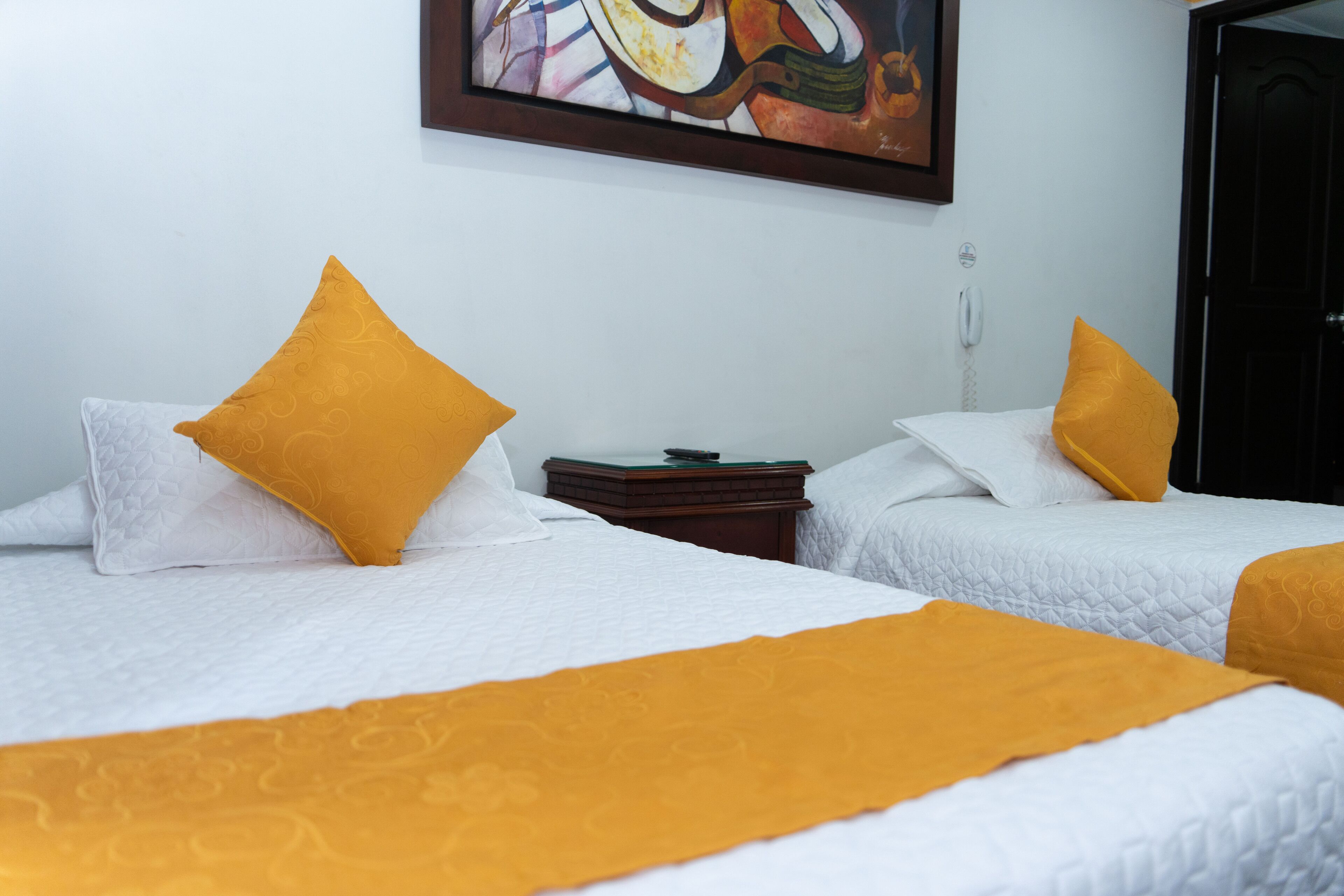 A hotel room with two beds, each with a white quilted bedspread and a single yellow pillow. There is a small wooden nightstand between the beds with a telephone and a small table. A painting is visible on the wall above the beds.