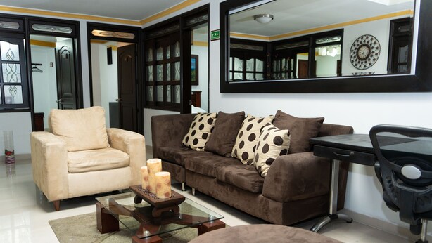 A living room with a beige armchair, a brown sofa with polka dot cushions, a glass coffee table with candles, and a black office chair. - Hotel Aires Del Quindio (Armenia)