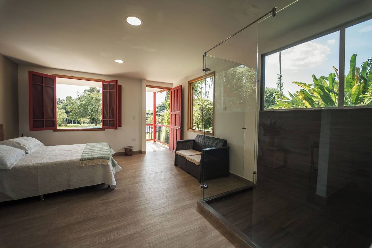 Un dormitorio con cama, sofá, separador de vidrio y ventanas amplias con vistas a la vegetación.
