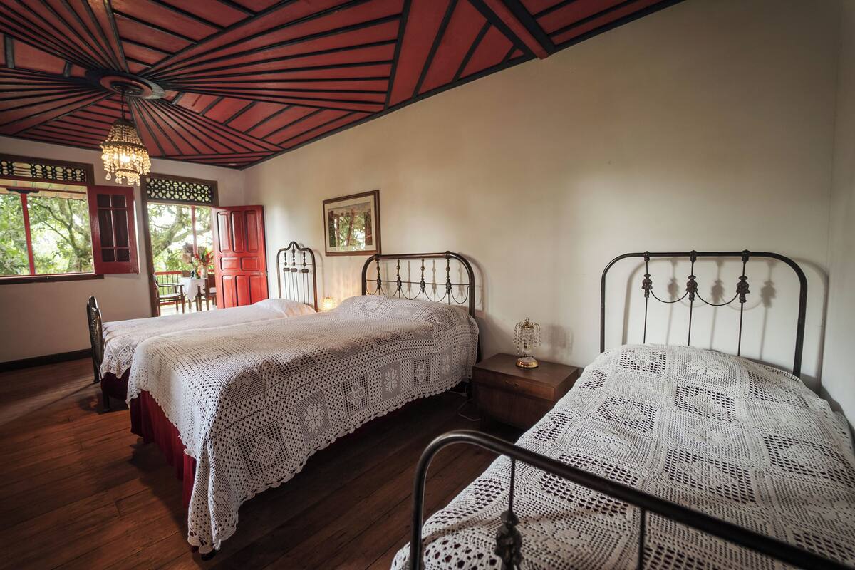 Una habitación con dos camas, piso de madera, un candelabro y una ventana con vista a árboles.