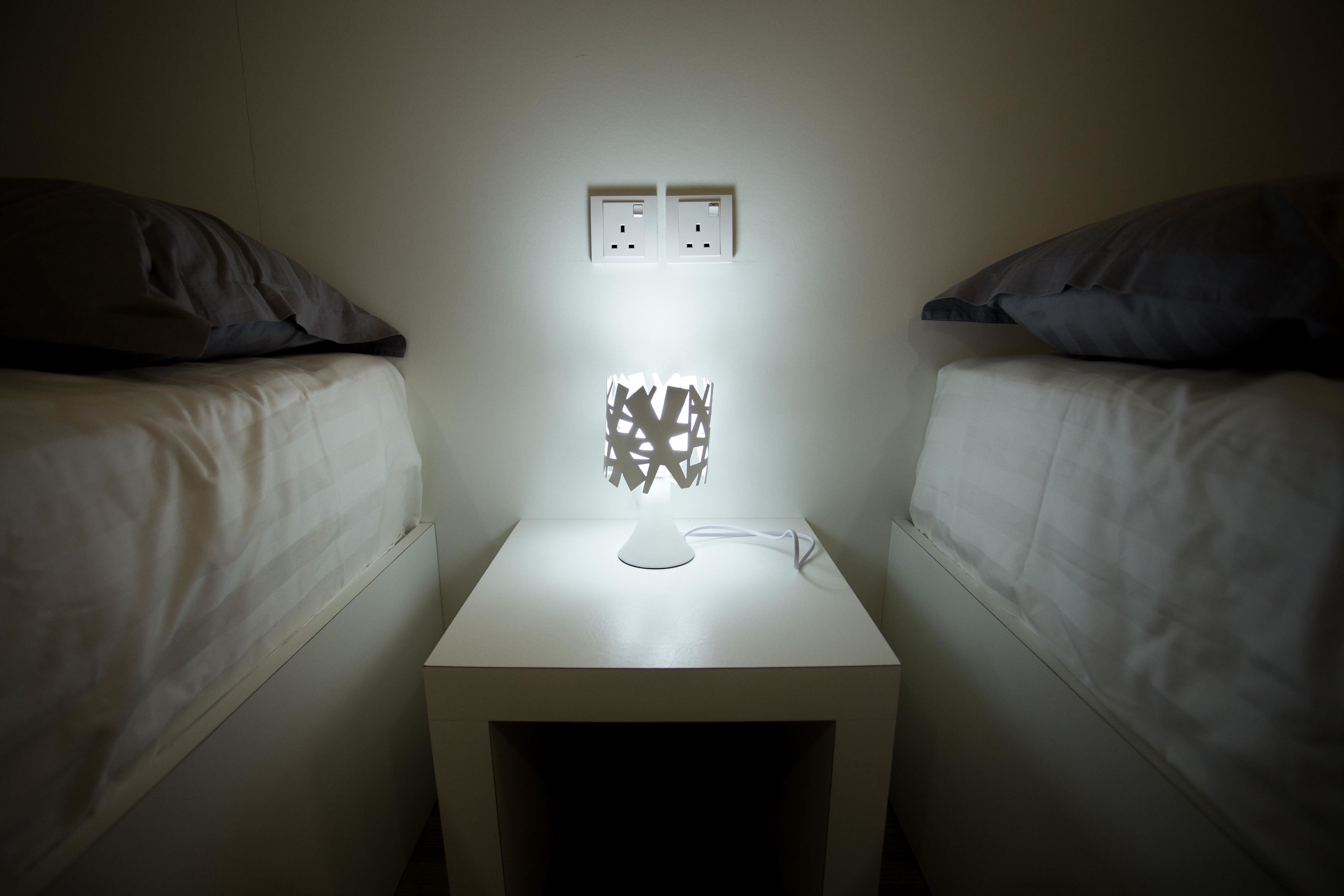 Una habitación pequeña y tenuemente iluminada con dos camas, una mesita de noche con una lámpara y dos enchufes en la pared.