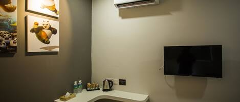 A hotel room with a flat-screen TV mounted on the wall, a white desk with a kettle and bottled water, and framed pictures of pandas on the wall.