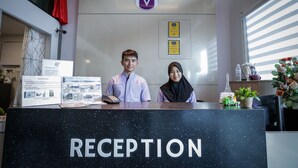 A reception desk with a sign reading "RECEPTION" and a logo above it. There are framed certificates on the wall behind the desk. - Merrinton Hotel (Temerloh)