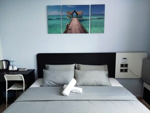 A hotel room with a bed, pillows, a desk, a chair, a nightstand, a lamp, and a wall-mounted power outlet. - Merrinton Hotel (Temerloh)